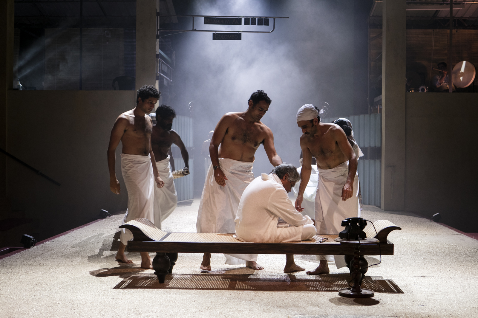 Five men in white cloths stand around an older man sitting on a bench, who is also dressed in white. The scene is dramatic, with mist and dim lighting, set on a stage with minimal props, including a black phone.