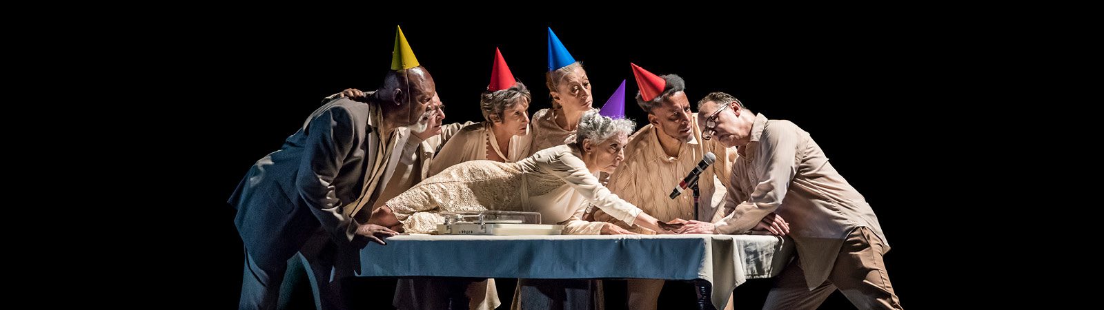 Six adults in party hats gather around a table, leaning in closely. A woman in white lies on the table reaching toward a cake, while the others watch or support her, all set against a black background.