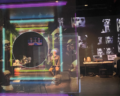 A person sits on stage surrounded by colorful neon-like rectangles, distorted faces projected behind, and people working at desks with computers and mannequin heads to the right.