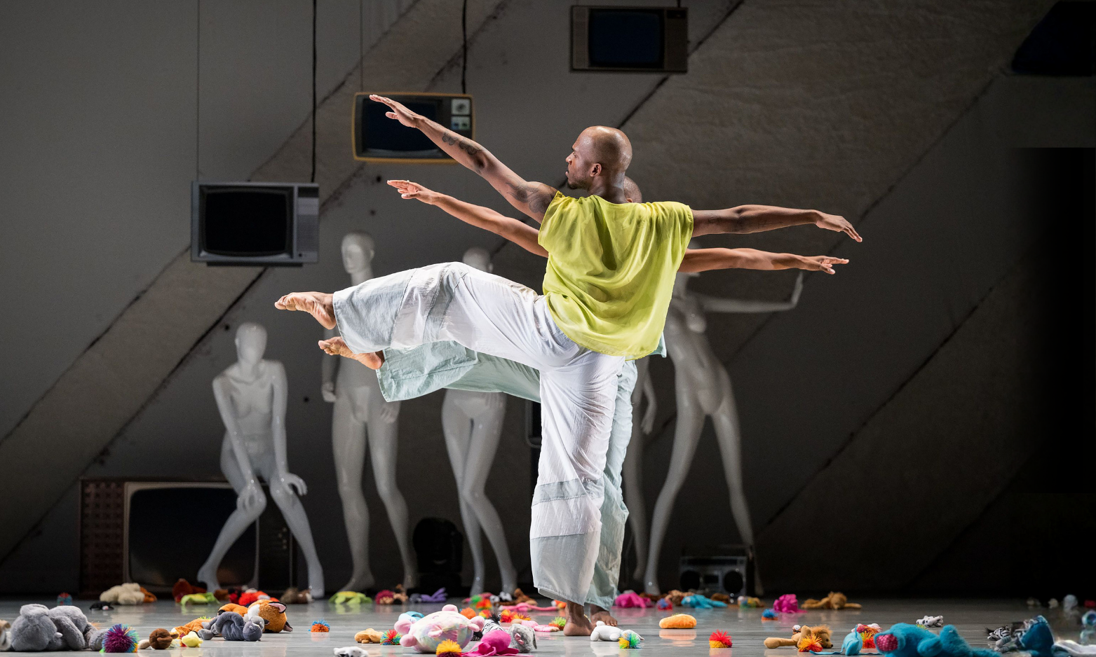 Two dancers in loose clothing pose balancing on one leg, arms extended. White mannequins, plush toys, and retro TVs surround them on a stage with a geometric backdrop.