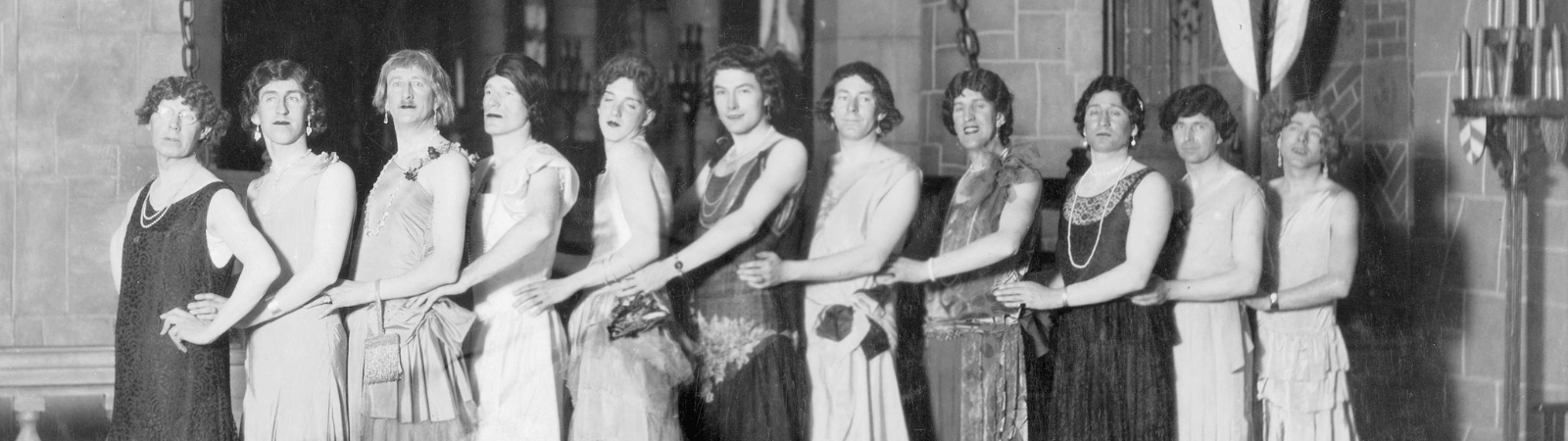 A black-and-white photo of ten people in dresses standing in a line, holding each other’s waists, and looking at the camera in what appears to be a festive or theatrical setting.