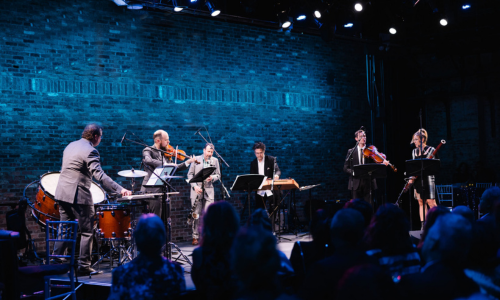 A small ensemble of musicians performs onstage with string and percussion instruments in front of an audience, with blue lighting and a brick wall backdrop.