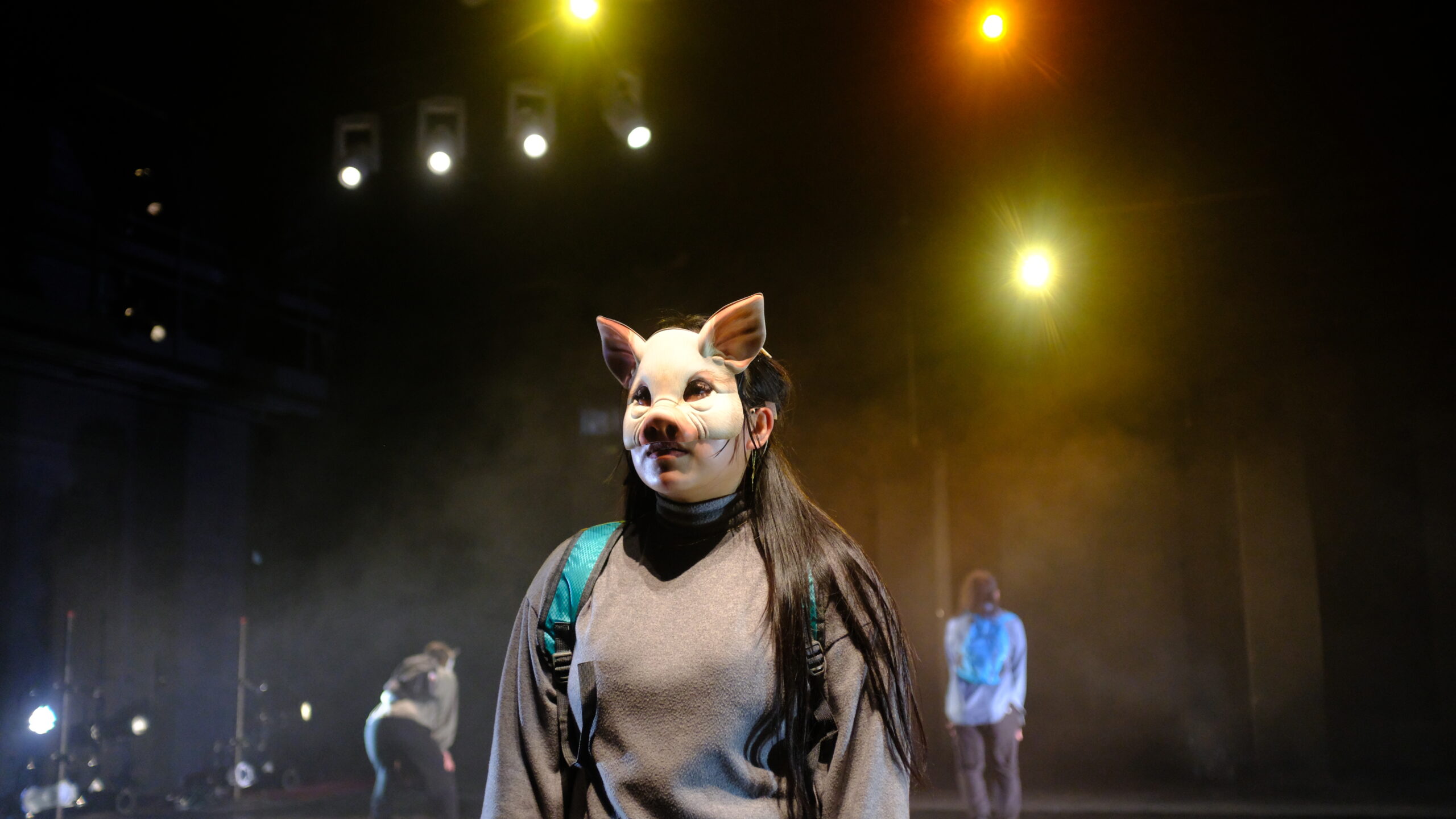 A person wearing a pig mask and gray sweater stands under stage lights, with others in the background and fog creating a dramatic atmosphere.