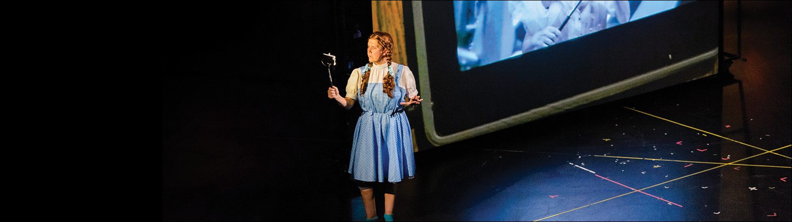 A performer in a blue gingham dress stands on stage holding a wand, with a projection screen behind her and colored tape marking the floor.