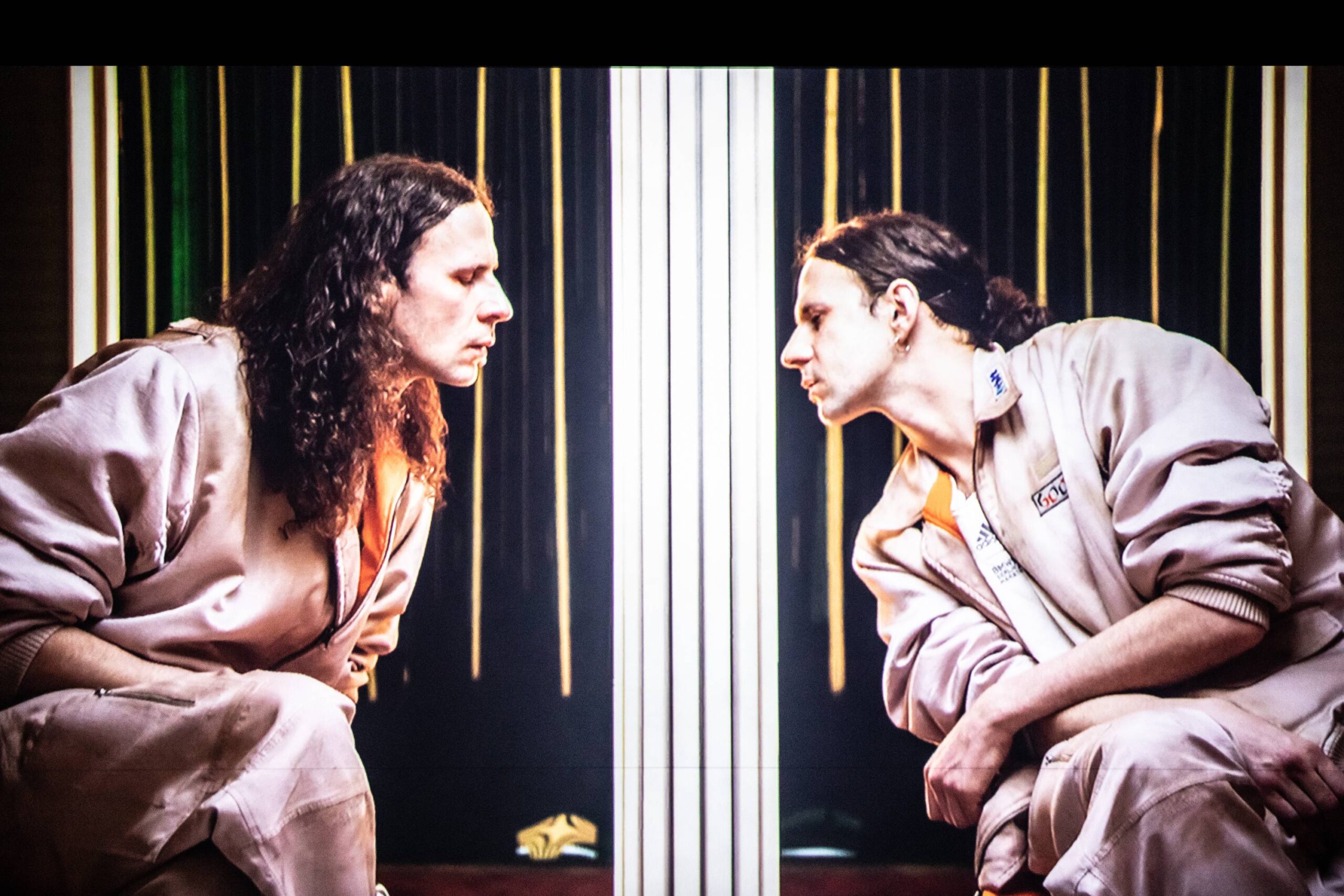 A person with long hair and a beige jacket sits squatting, gazing intently at their reflection in a mirror, with vertical lines in the background.