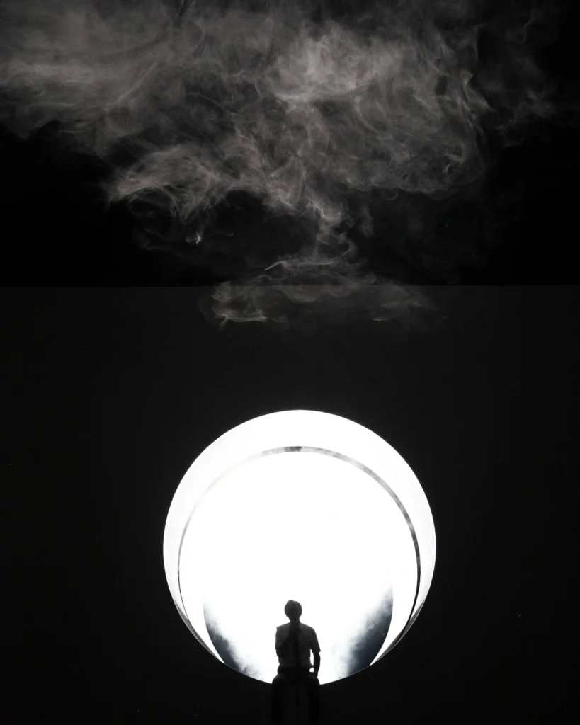 A silhouette of a person sits in front of a large, bright circular light with swirling white smoke above against a dark background.