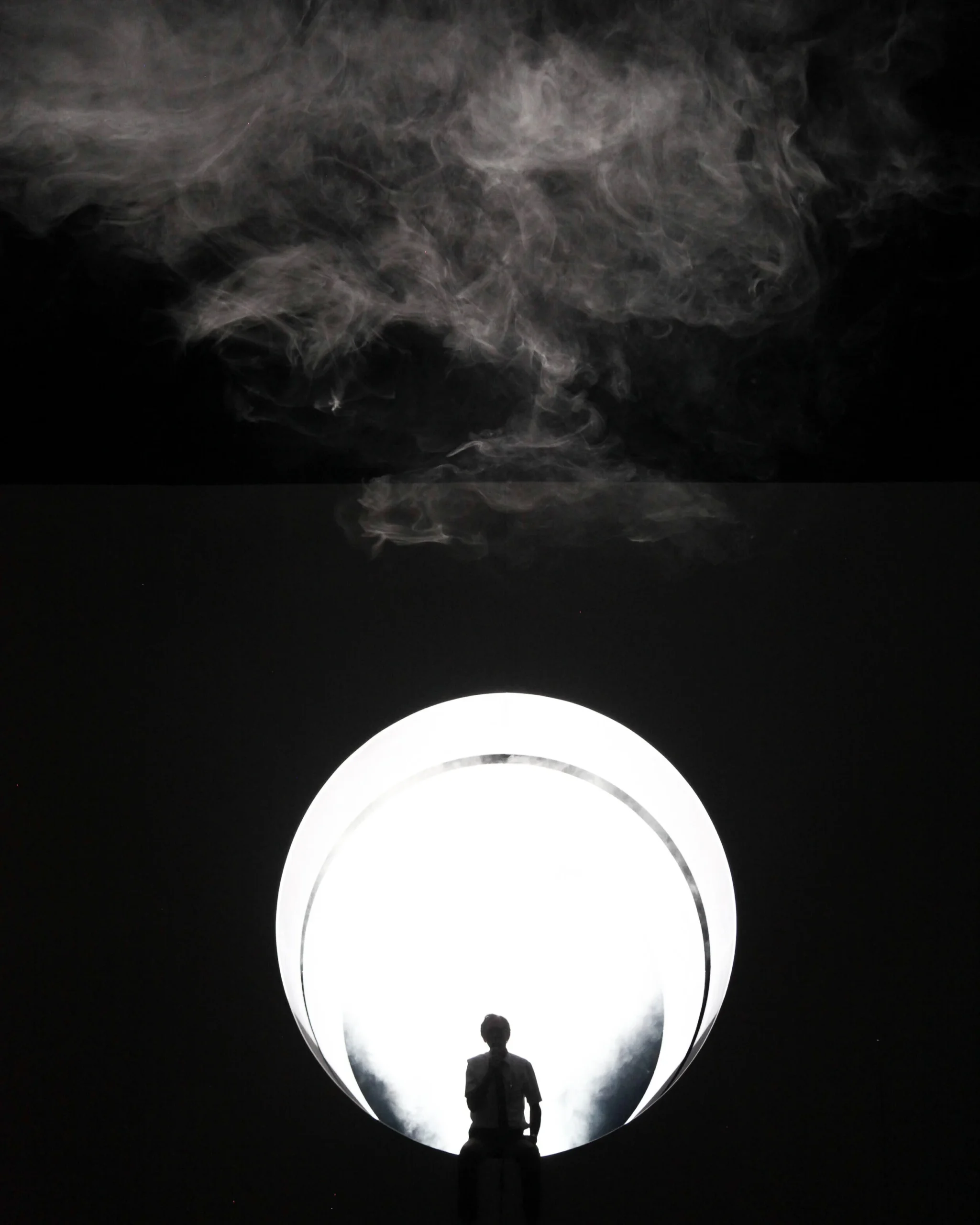 A silhouette of a person sits in front of a large, bright circular light with swirling white smoke above against a dark background.