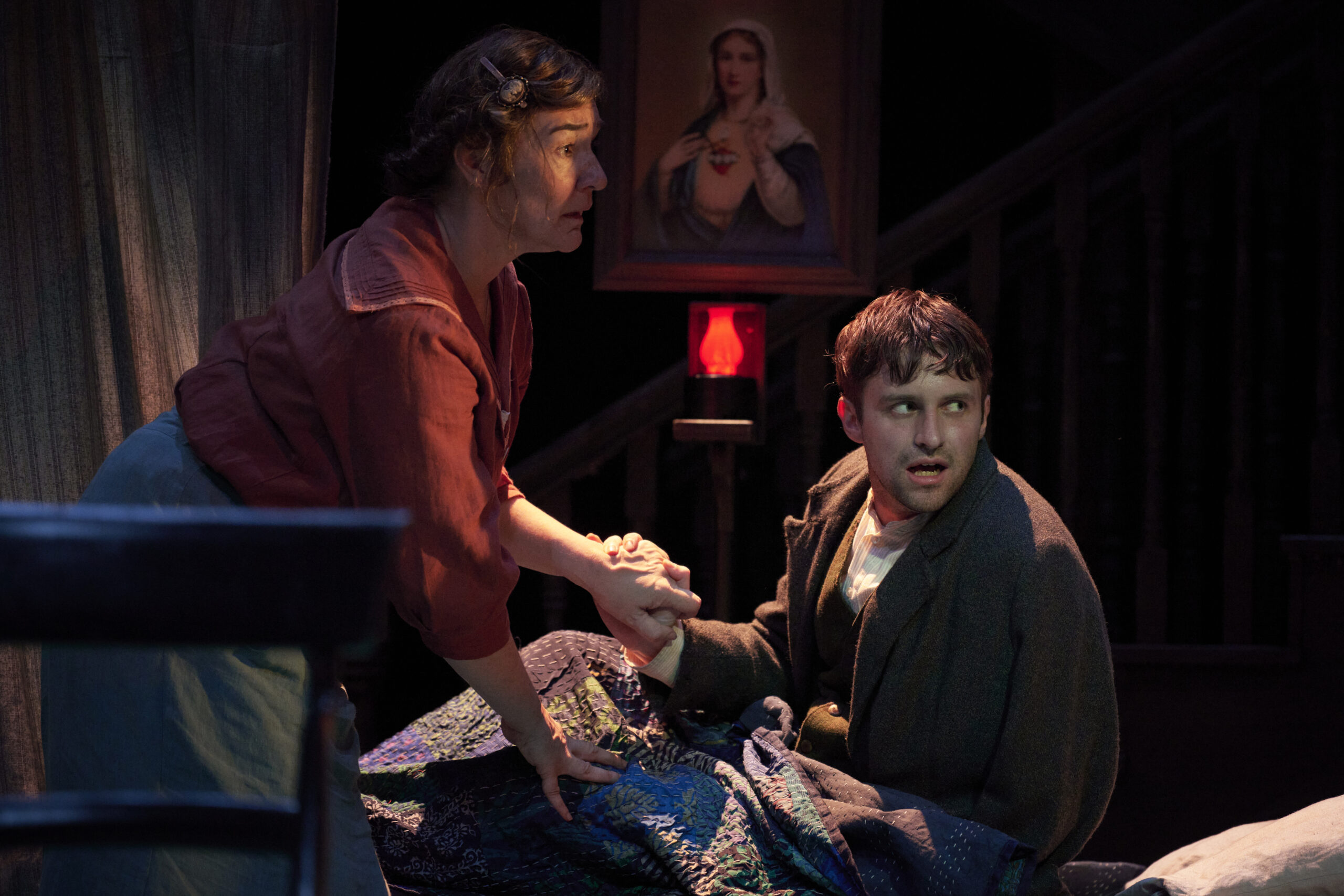 A woman holds a man’s hand as he sits up in bed, looking worried. A painting of the Virgin Mary and a red lamp are visible in the dimly lit background.