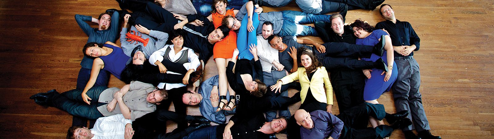 A group of people lies on a wooden floor in a playful, overlapping arrangement, forming a loose circle and smiling up at the camera.