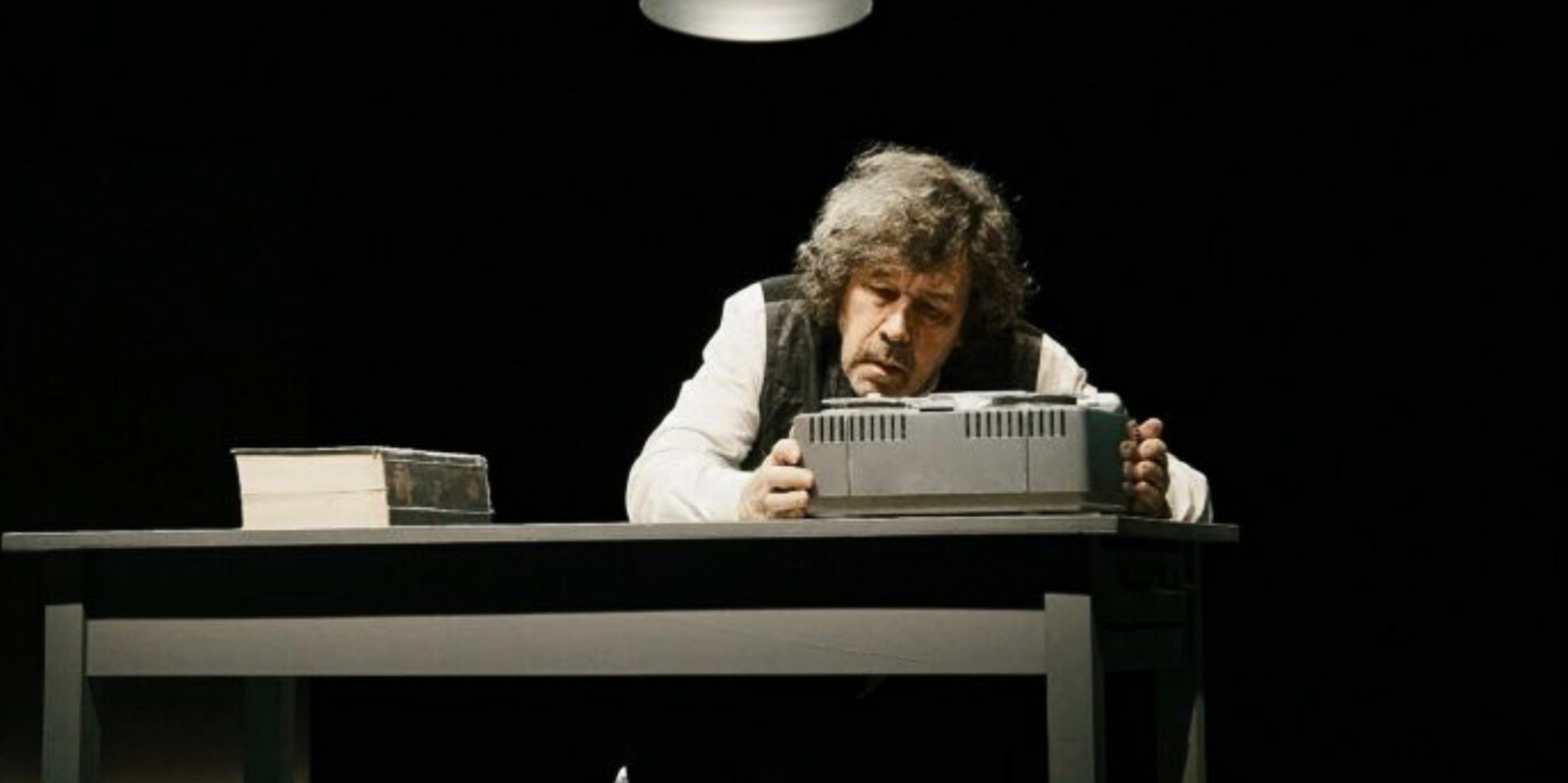 A man sits alone at a desk under a spotlight, examining a typewriter, with a closed book beside him. The background is dark and empty.