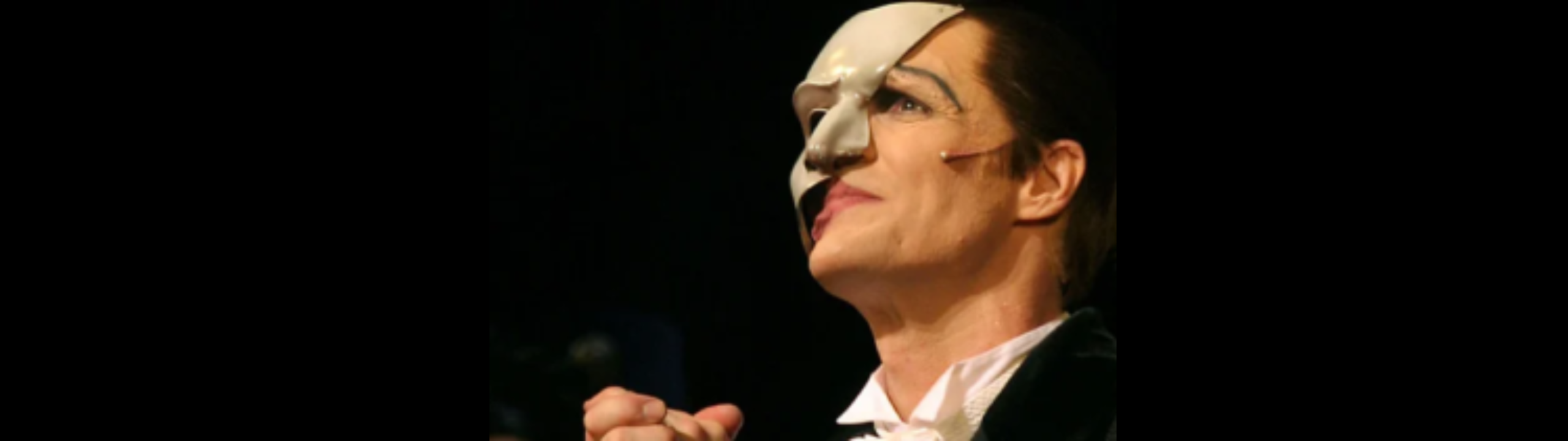A person wearing a white half-face mask and formal costume is on stage, with a dramatic expression and clenched hand, reminiscent of “The Phantom of the Opera.”.