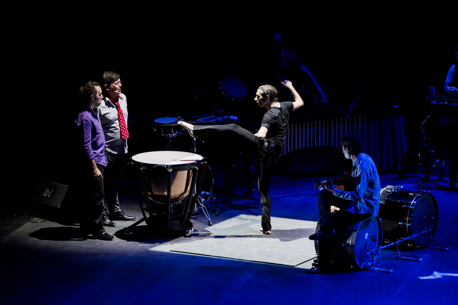 Four performers are on a dimly lit stage. One person stands on one leg, kicking high near two drums, while the others watch. Two are standing, and two are seated near percussion instruments under spotlights.