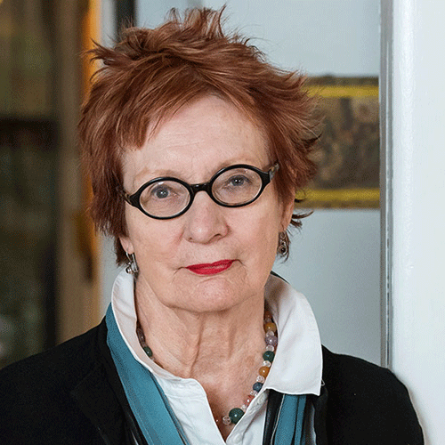An older woman with short, tousled reddish hair, black round glasses, and a serious expression, wearing a white collared shirt, beads, and a black jacket, stands indoors against a blurred background.