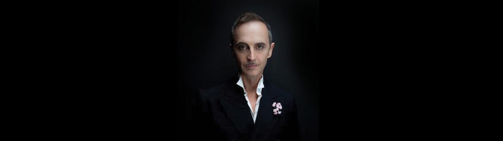 A person with short hair and a mustache, wearing a dark suit jacket with a white shirt and a pink flower on the lapel, sits against a black background, looking directly at the camera.