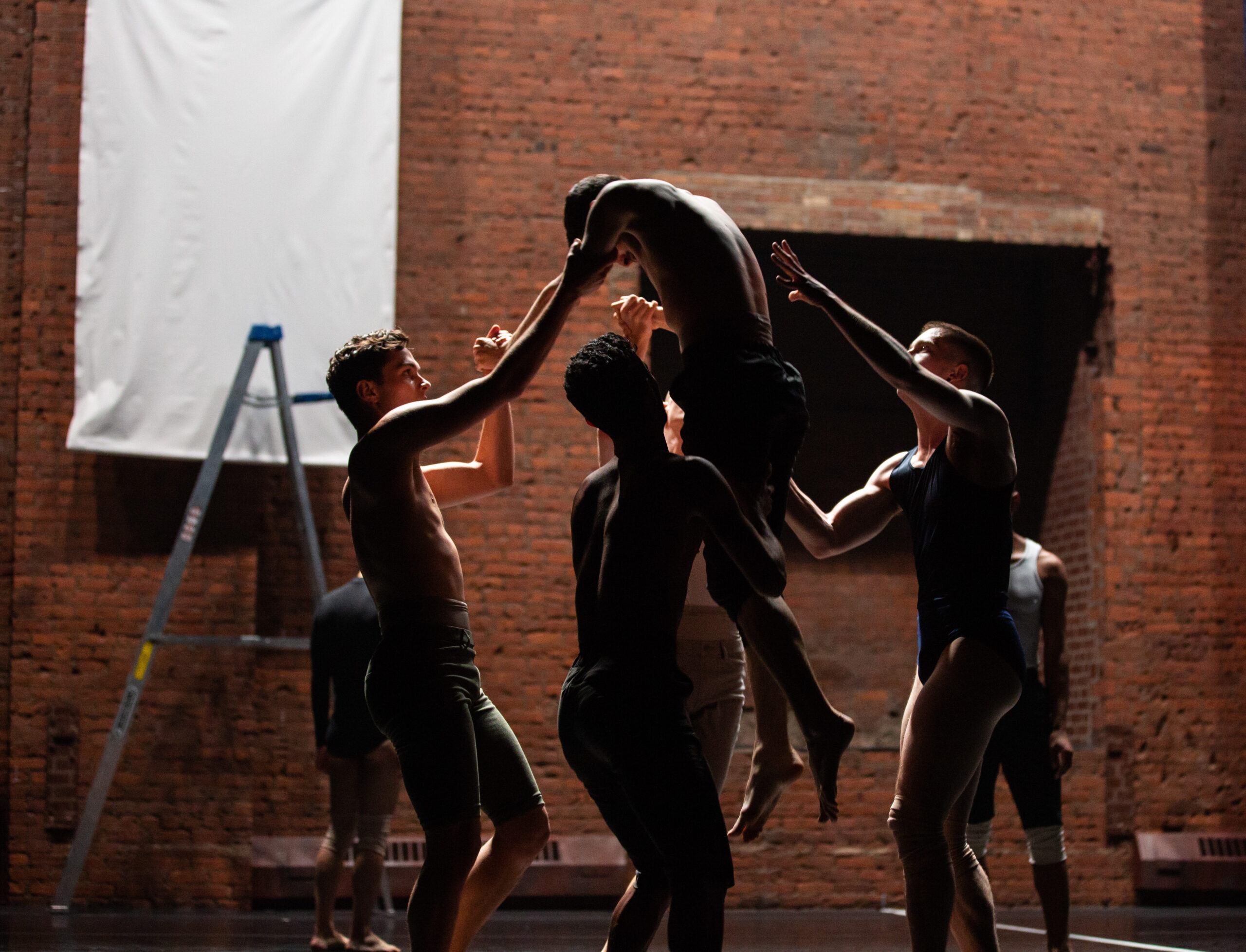 Four dancers lift another dancer in a dimly lit studio with brick walls and a ladder in the background.