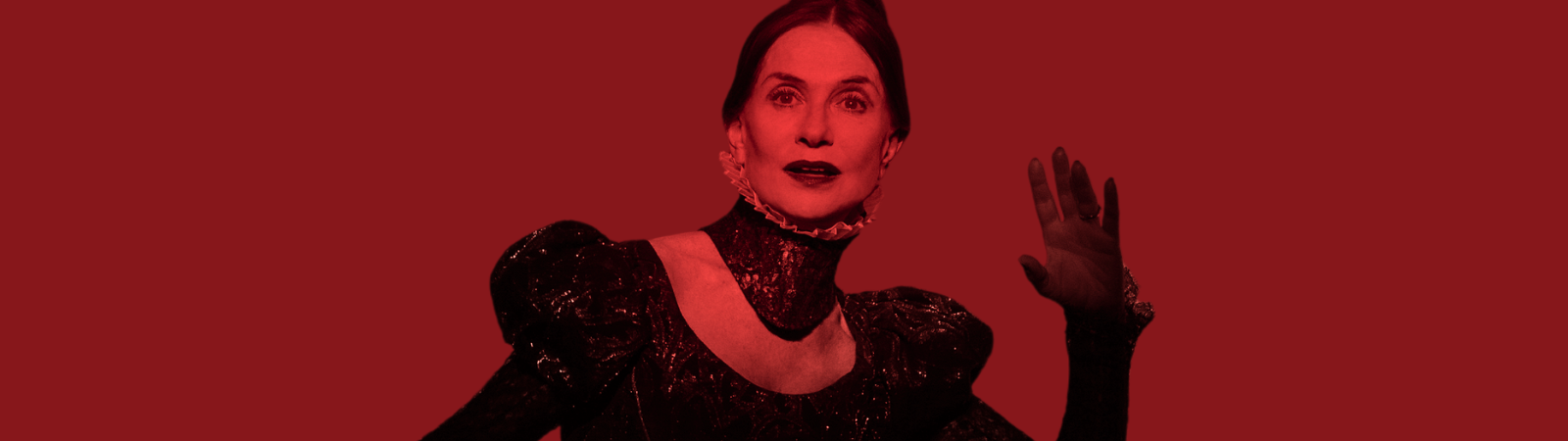 Person in a high-collared, dark, ornate outfit poses with one hand raised against a solid red background.