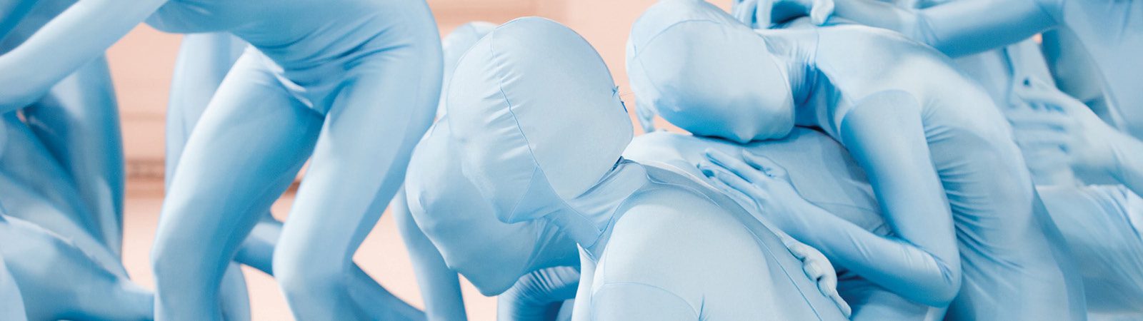 Several people wearing tight, full-body blue suits are intertwined in a dynamic, abstract pose, creating a surreal and sculptural visual effect.