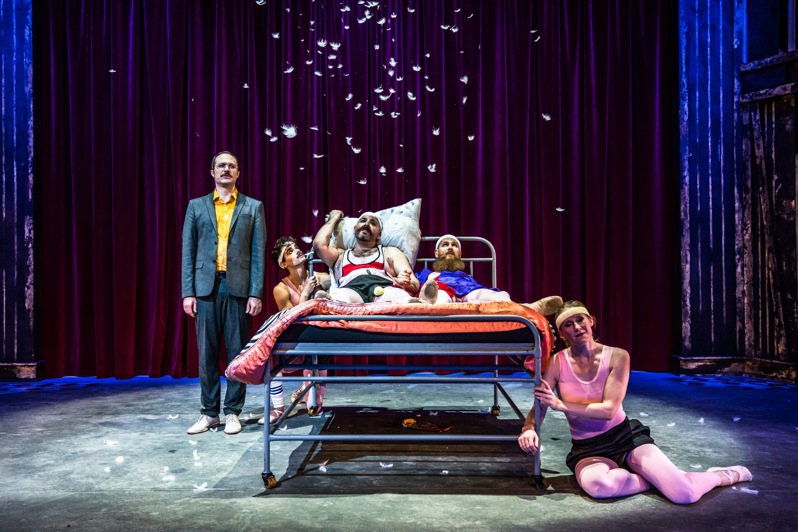 Five actors in theatrical costumes surround a hospital bed on stage, with feathers falling in midair. A red curtain hangs in the background, and dramatic lighting highlights the scene’s surreal atmosphere.