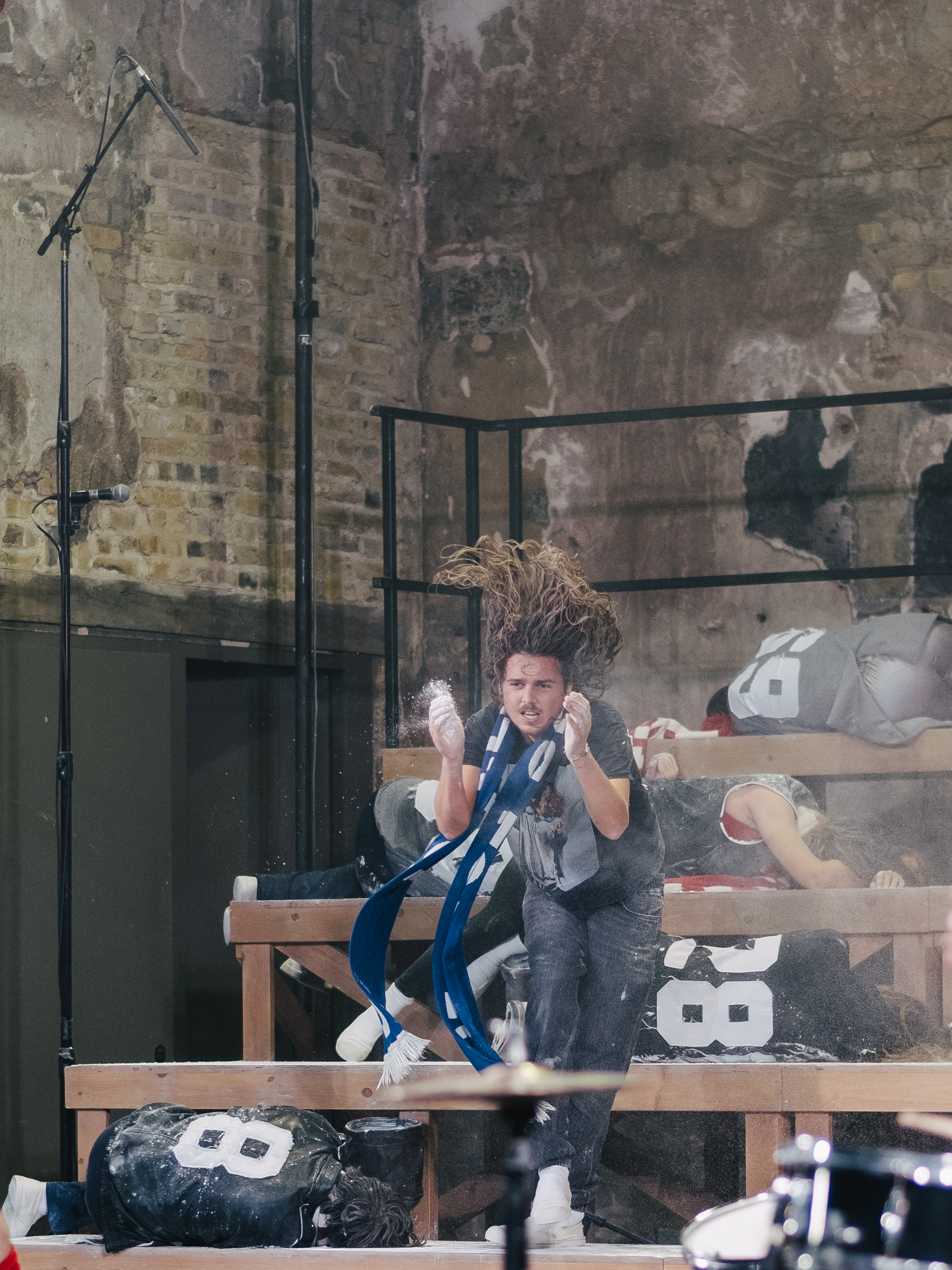 A person with long hair jumps energetically in front of a row of benches with mannequins in sports jerseys. Dust or chalk fills the air in an indoor, rustic setting with exposed brick walls.