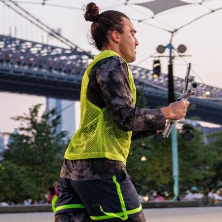 A person wearing a yellow vest and patterned athletic clothes runs outdoors, holding a handheld gimbal. A bridge and trees are visible in the background.