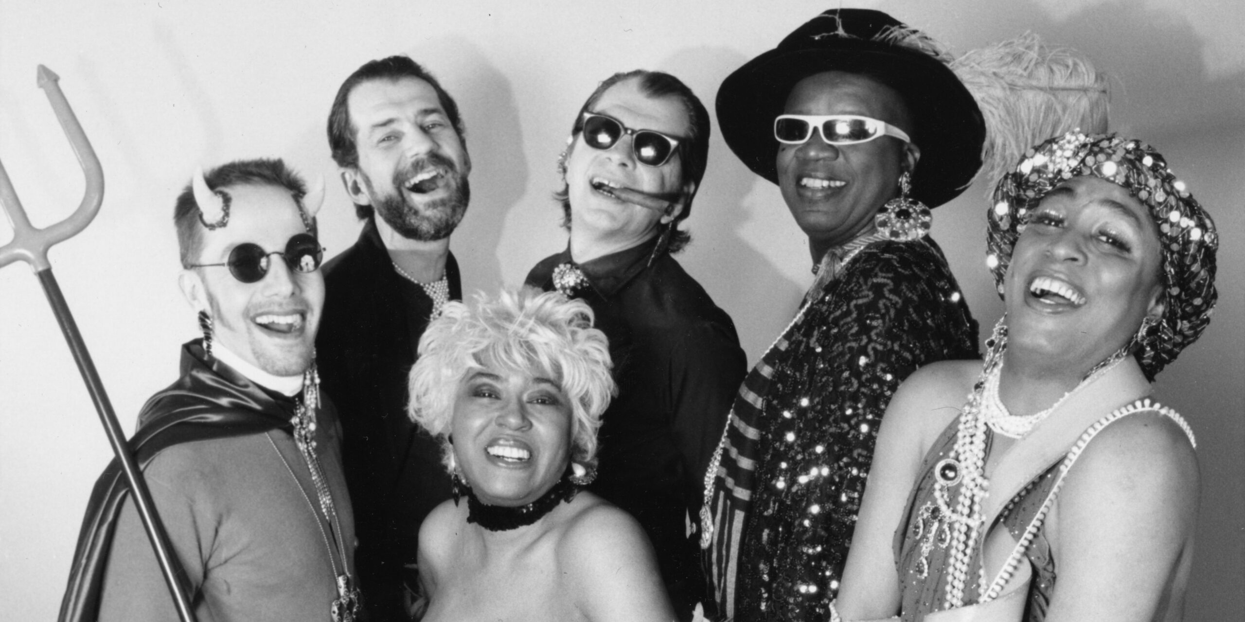 Six people dressed in festive, glamorous costumes and accessories, including a trident, sunglasses, horns, and sequins, pose together smiling and laughing in a black and white photo.