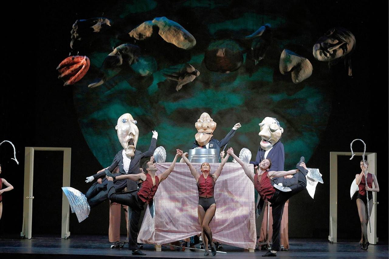 Performers with large, exaggerated puppets and masks dance on stage around a table, while floating heads and fish hang above. Two dancers strike high kicks beside a checkered tablecloth in a theatrical, surreal scene.
