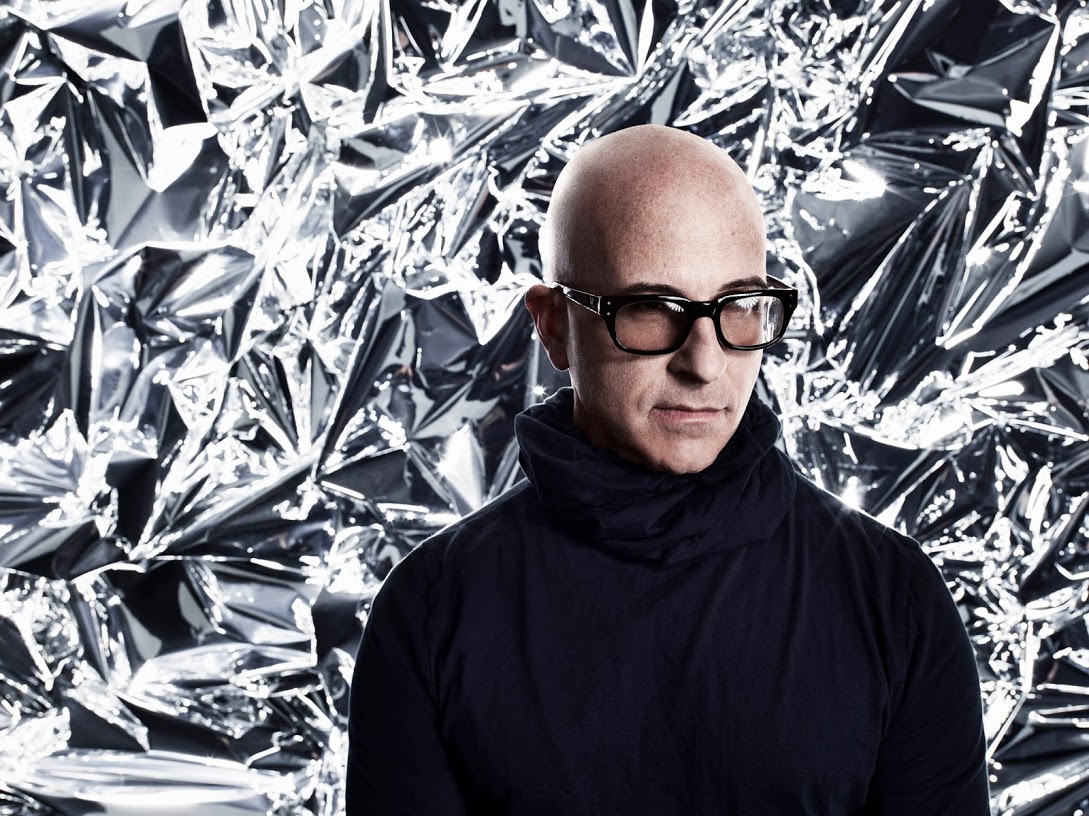 A bald person wearing black glasses and a dark turtleneck stands in front of a shiny, crumpled metallic foil background, looking slightly downward.