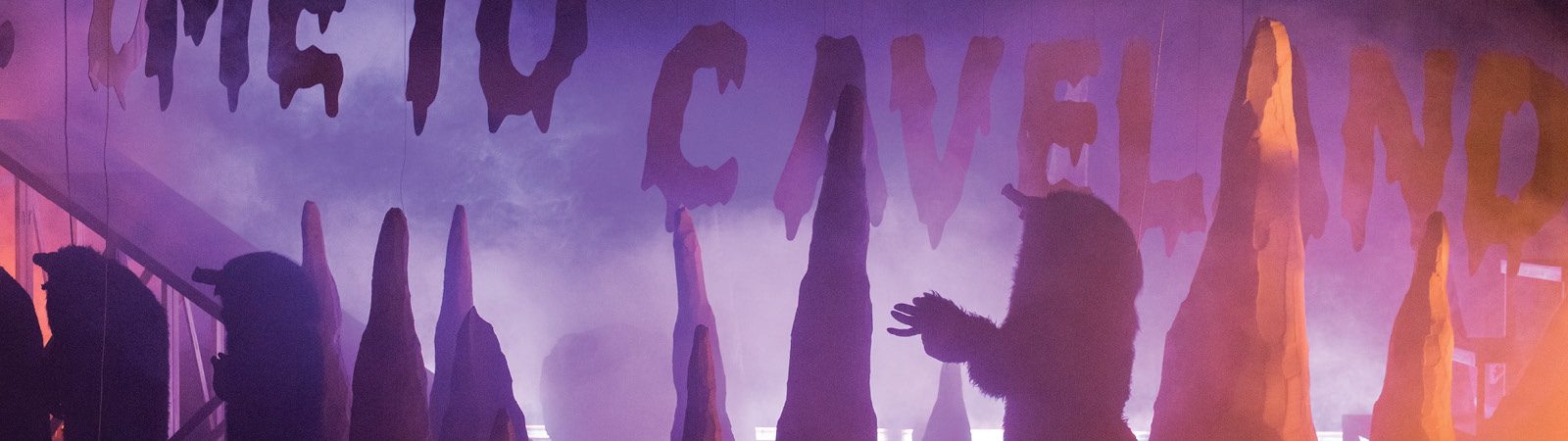 Silhouetted people walk beneath a sign reading “CAVELAND” in jagged letters, with hanging decorations and purple fog creating a mysterious, cave-like atmosphere.