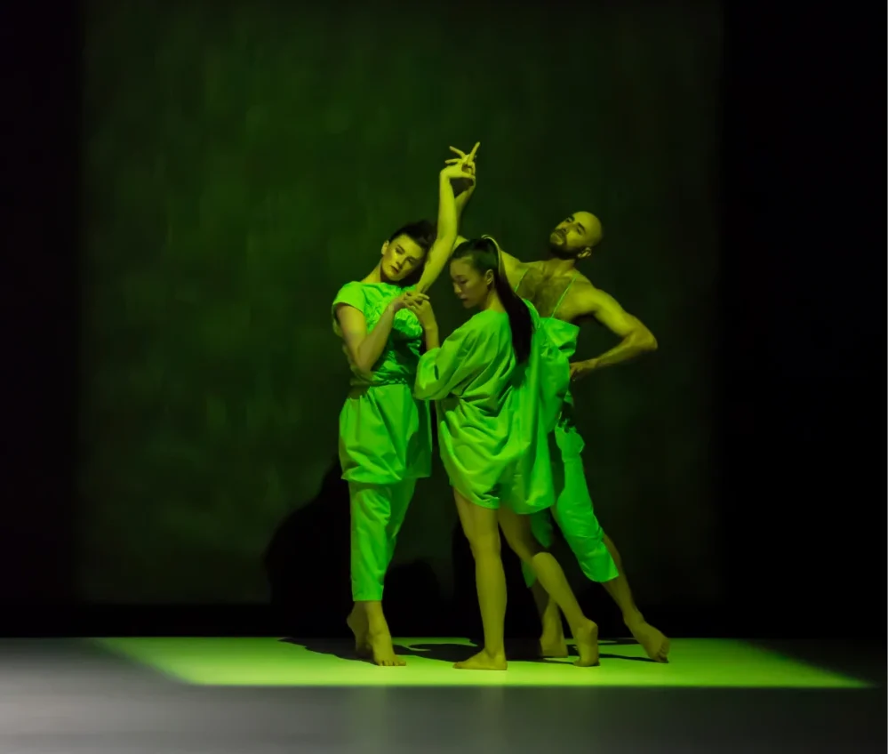 Three dancers in green costumes pose closely together on stage under dramatic green lighting, creating an artistic and expressive scene.