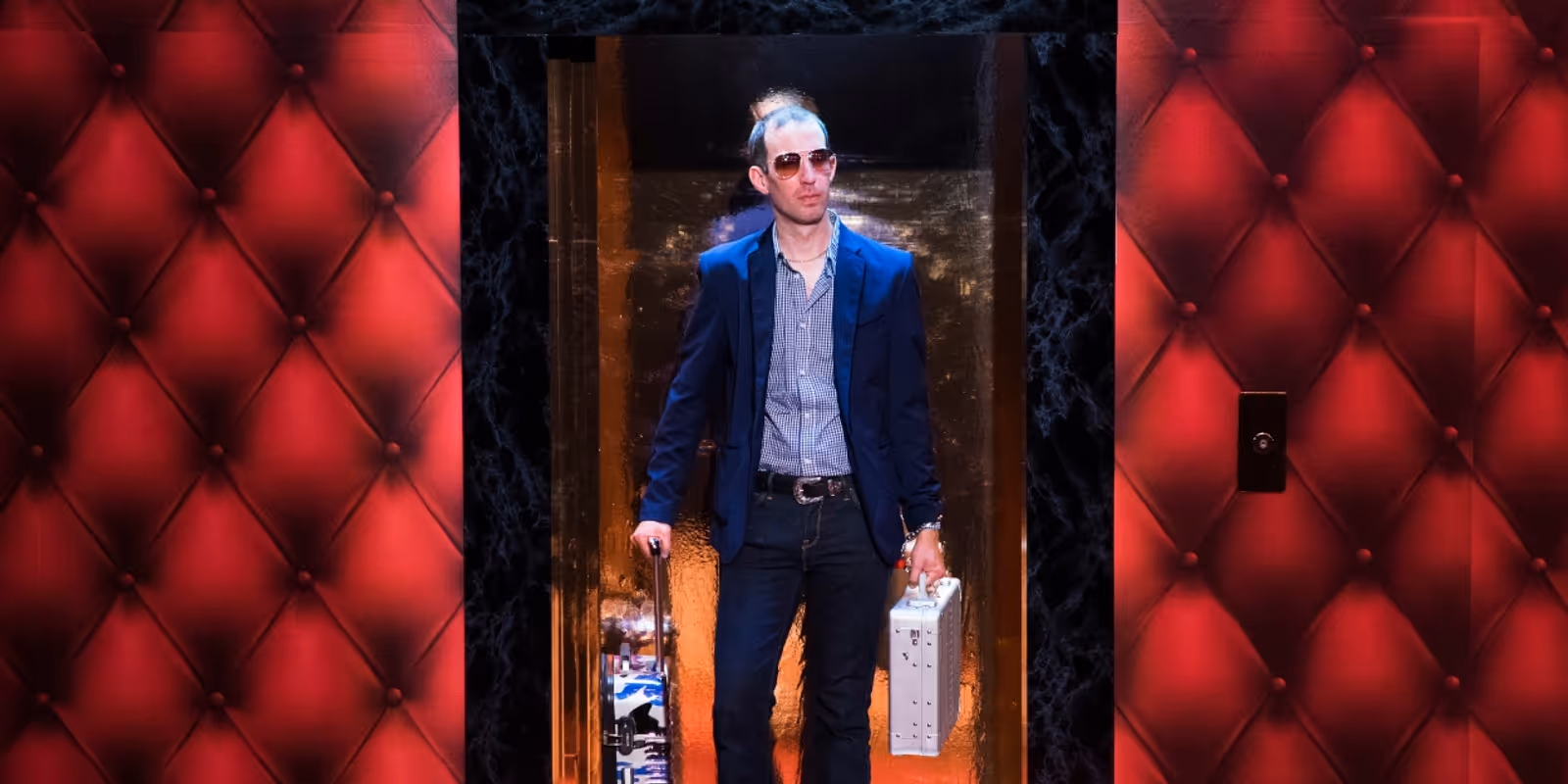 A man in sunglasses and a blazer stands in an elevator holding a metallic briefcase, with red padded walls on either side of the elevator door.