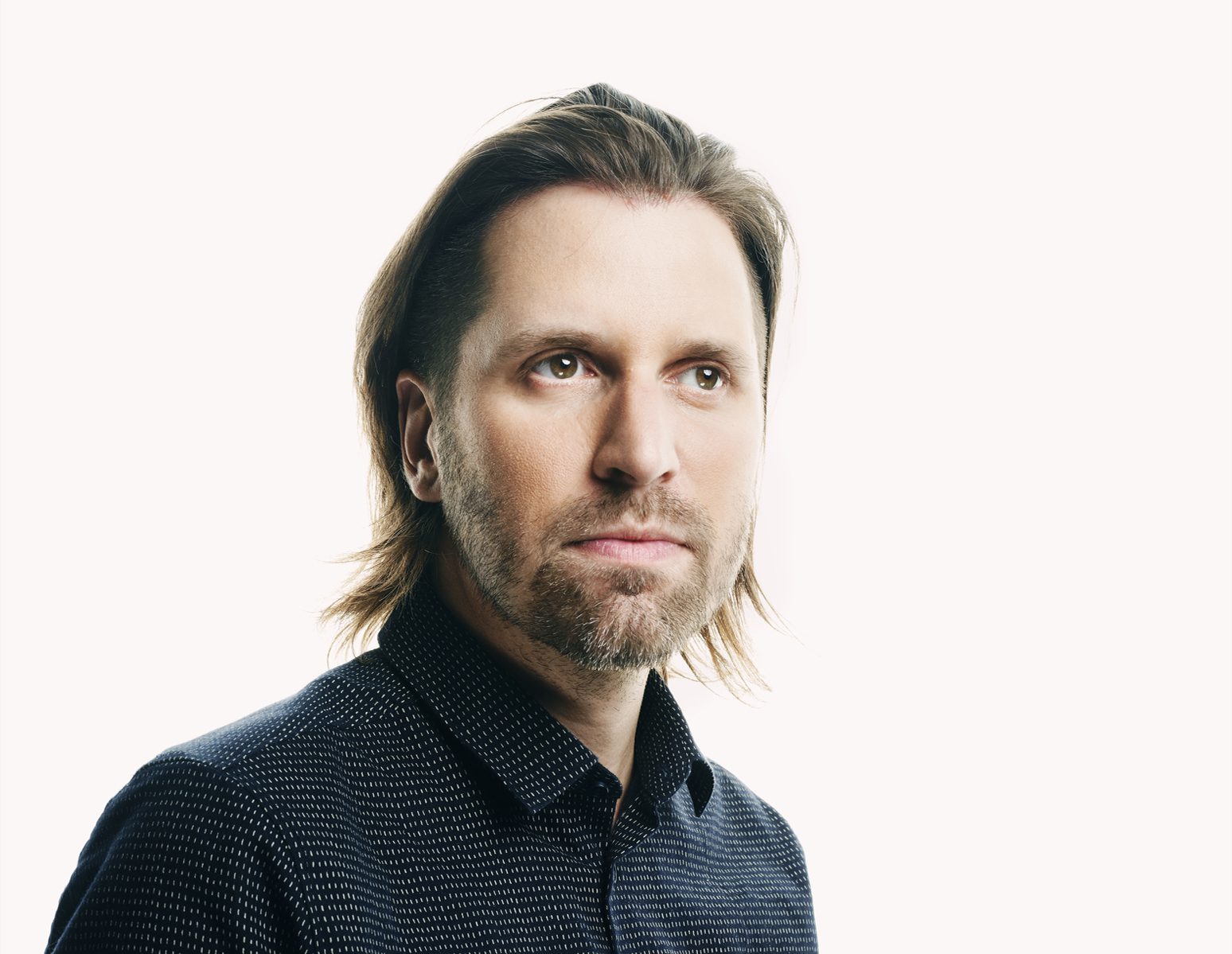 A man with shoulder-length brown hair and a beard wearing a dark collared shirt looks slightly upward against a plain white background.