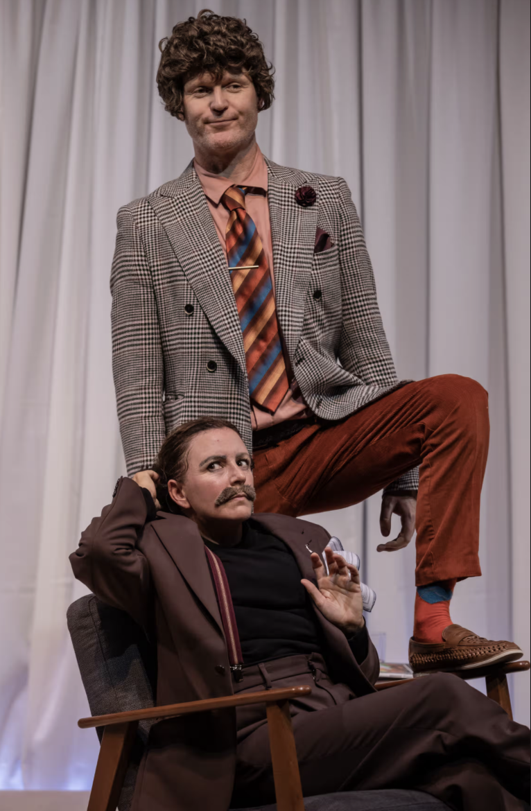 Two people in vintage clothes pose dramatically; one stands with a foot on a chair, smirking, while the other sits below, looking up with a fake mustache. White curtain background.
