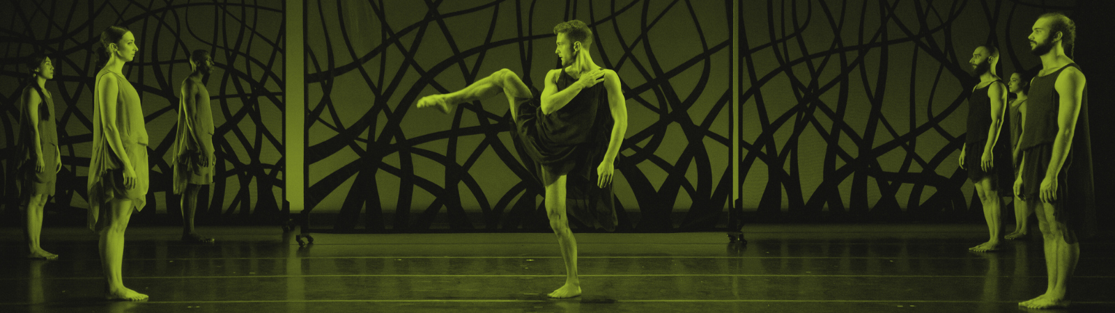 Several dancers stand on stage, barefoot, wearing flowy costumes. One dancer in the center raises a leg and arm dramatically. The backdrop features an abstract, tangled line pattern. The image has a green-yellow tint.