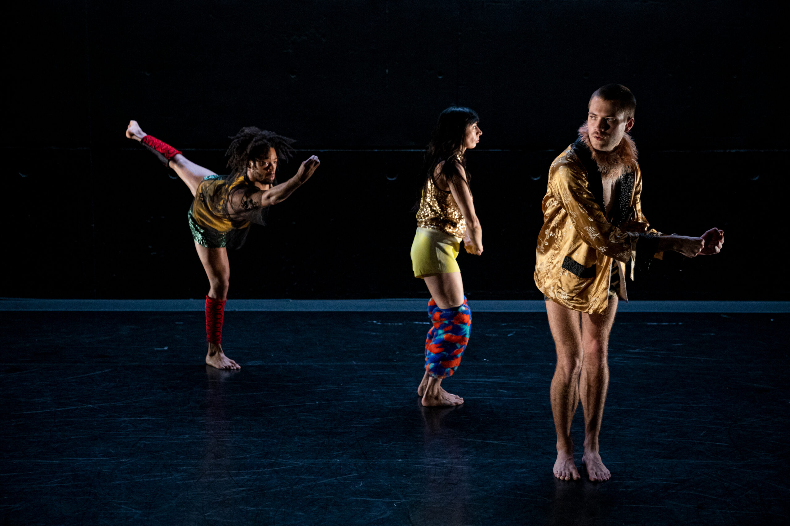Three dancers perform on a dark stage, wearing gold and colorful costumes. One dancer kicks a leg back, another stands in profile, and the third faces forward, each striking a unique pose.