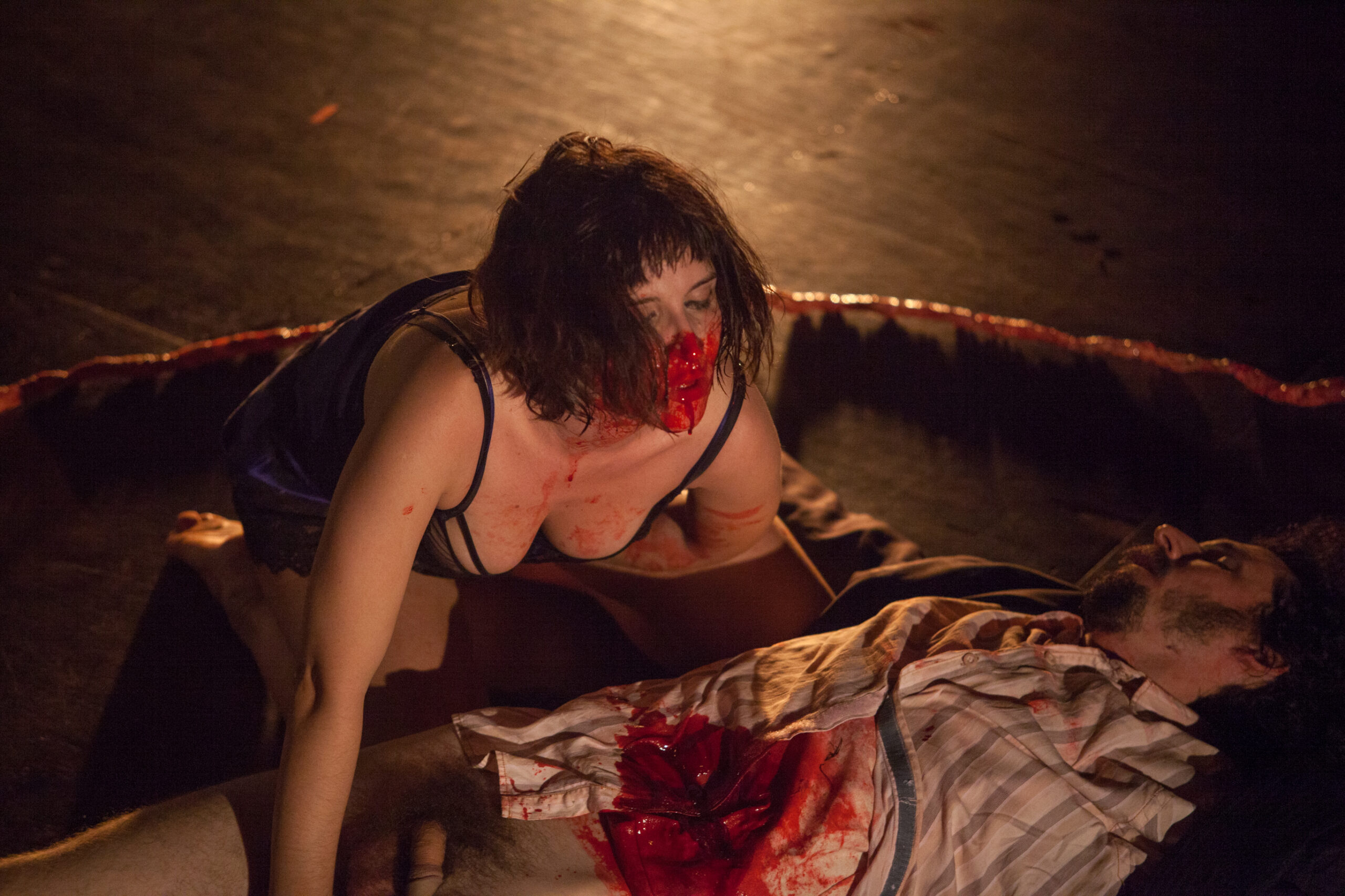 A woman with blood on her face crouches over a man lying on the ground, his shirt open and covered in fake blood, in a dark, dramatic setting.
