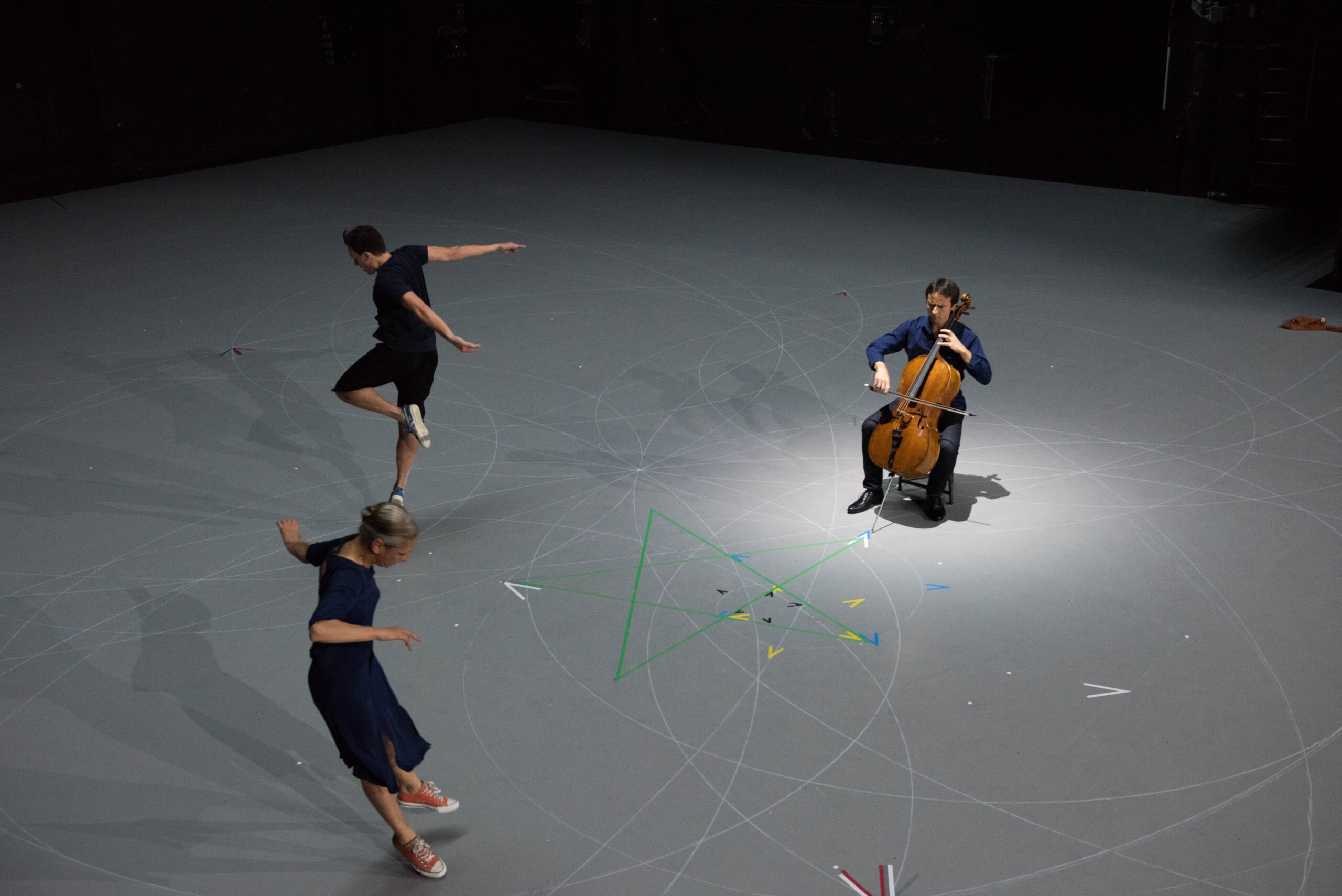 A cellist plays while two dancers move around him on a gray stage marked with intersecting lines and colored triangles, under soft lighting.