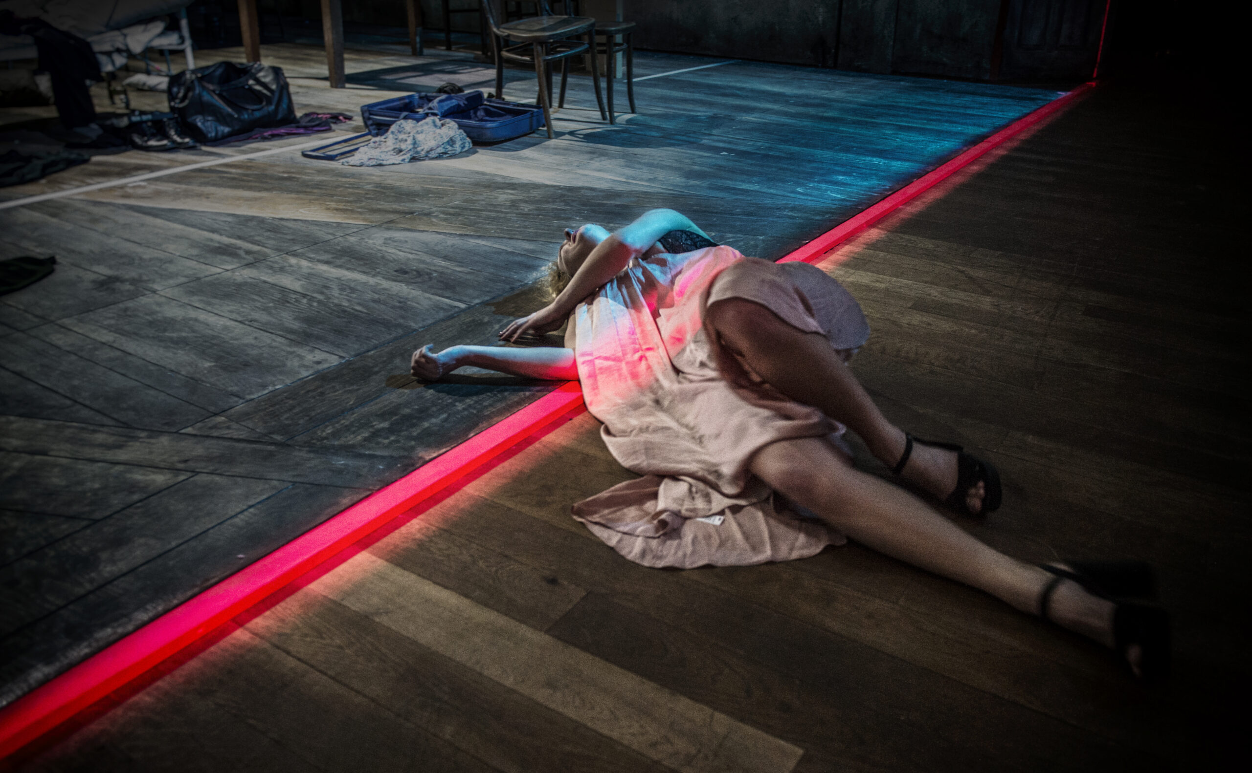 A woman in a light dress lies on a wooden floor near a glowing red line, with chairs and scattered belongings in the background.