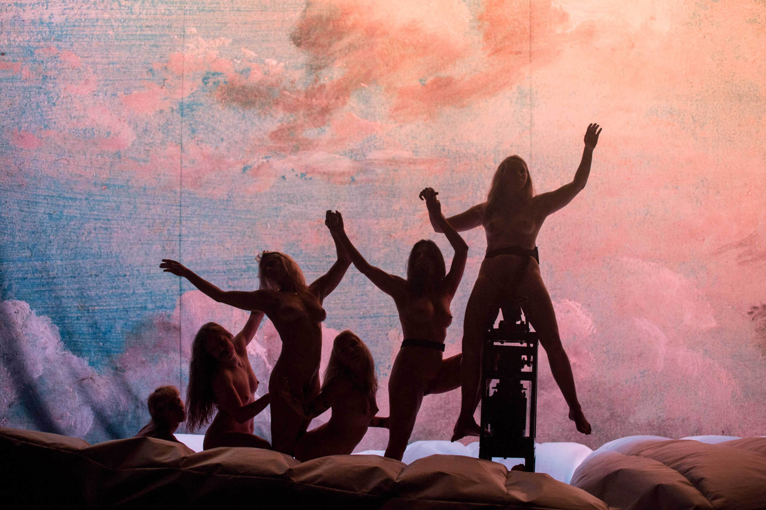 Six people pose in playful, dynamic stances, silhouetted against a pastel sky backdrop with clouds. The lighting emphasizes their outlines and expressive gestures.