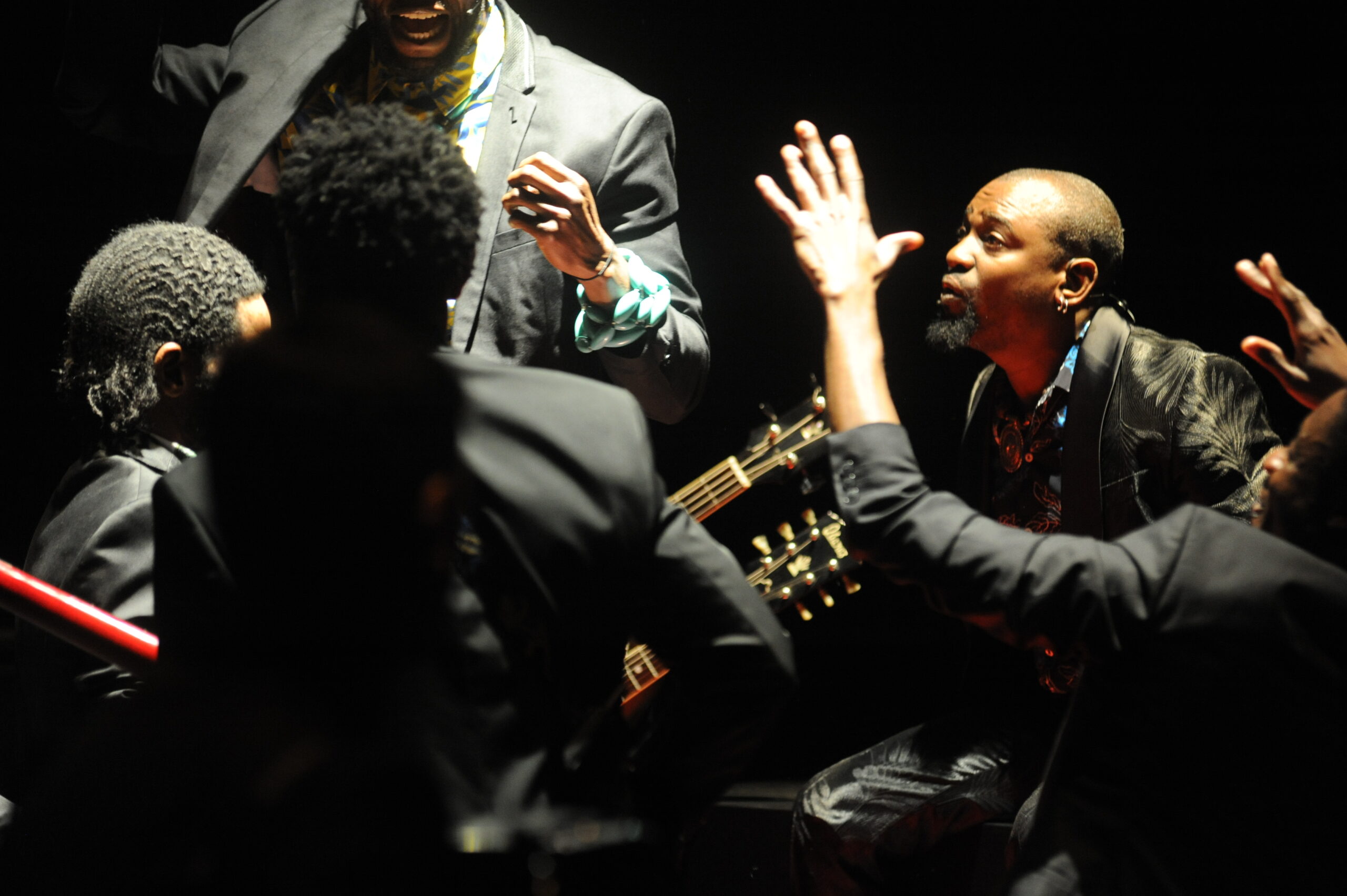Five men in dark suits gathered closely under dramatic lighting, some with hands raised in expressive gestures. A guitar is partially visible among them, suggesting a musical or theatrical performance.