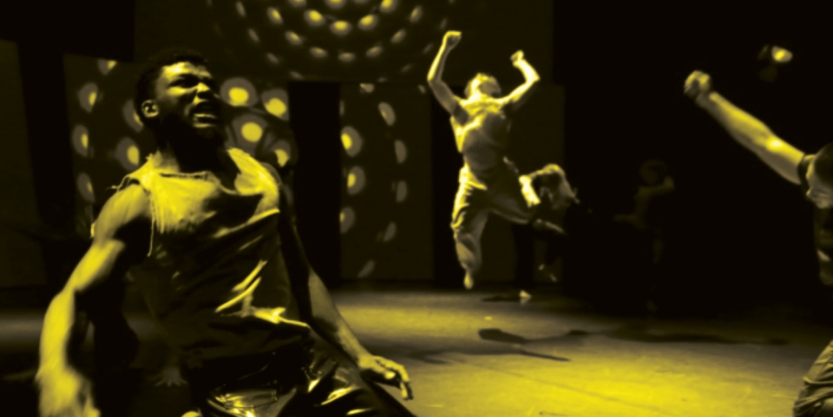 Dancers perform energetically on stage with dramatic lighting and circular light patterns in the background, some leaping into the air.