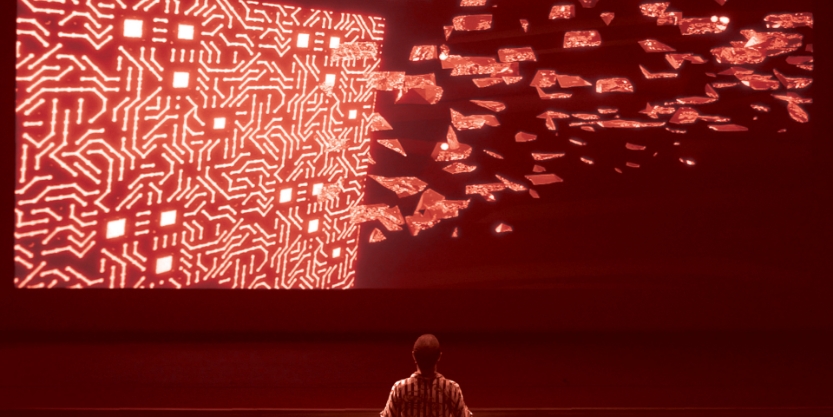 A person stands facing a large red digital screen with circuit-like patterns, watching as glowing fragments break away and float outward.