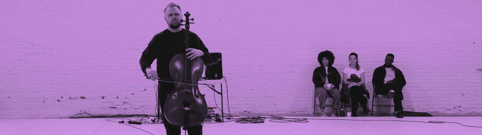 A person plays the cello in the foreground while three people sit against a brick wall in the background. The image has a purple tint.