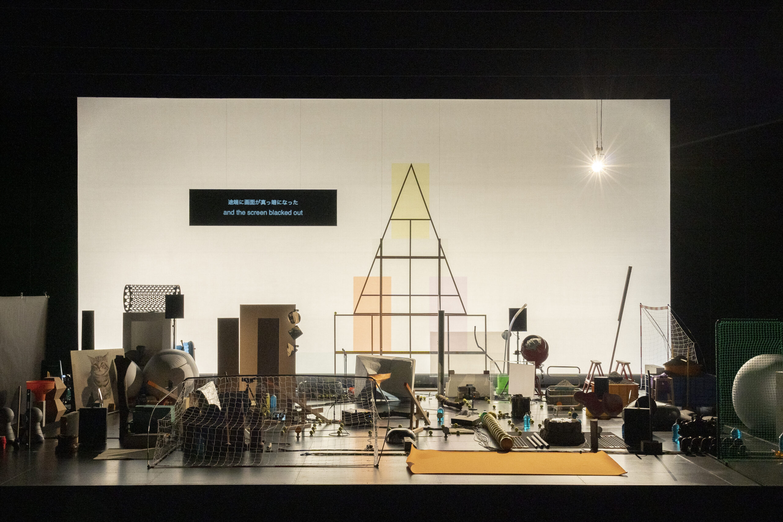 A brightly lit stage features various scattered objects, nets, boxes, and geometric frames. A screen above displays text in Japanese and English: “and the screen blacked out.”.