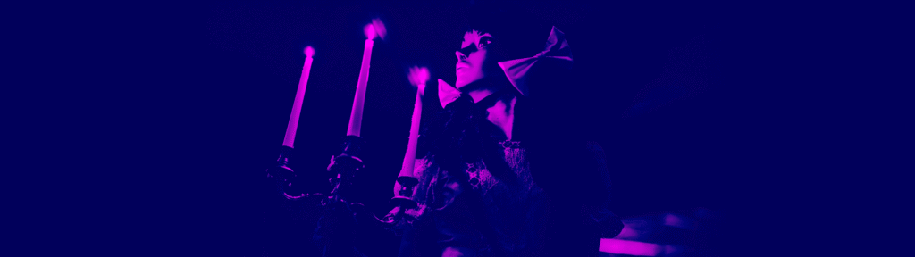 A person in a dramatic cape stands beside a candelabra with three lit candles, illuminated in deep purple and magenta lighting.