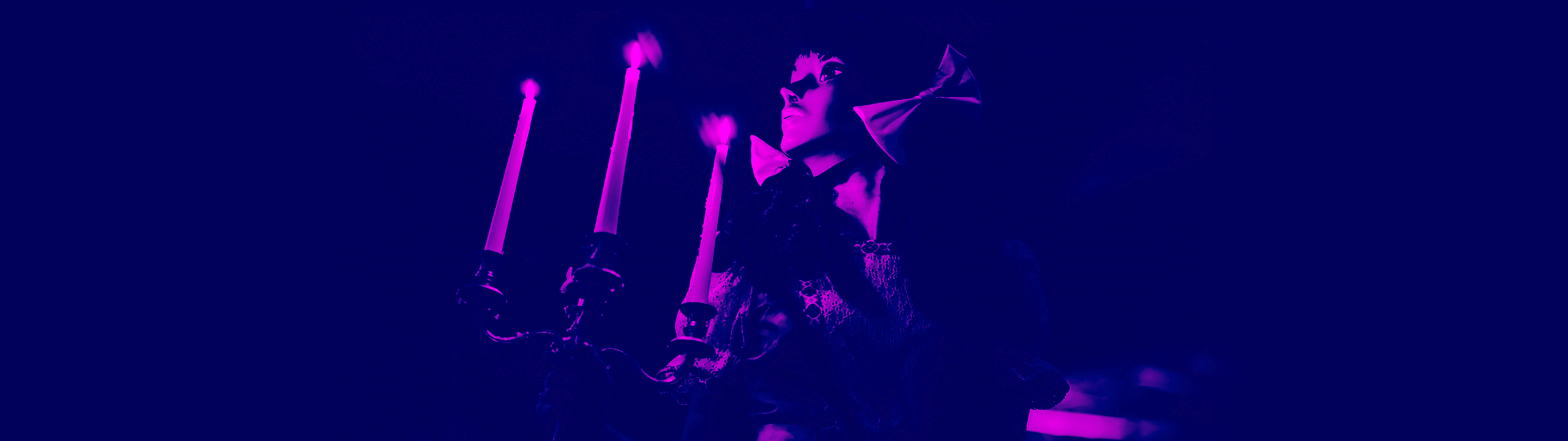 A person in a dramatic cape stands beside a candelabra with three lit candles, illuminated in deep purple and magenta lighting.