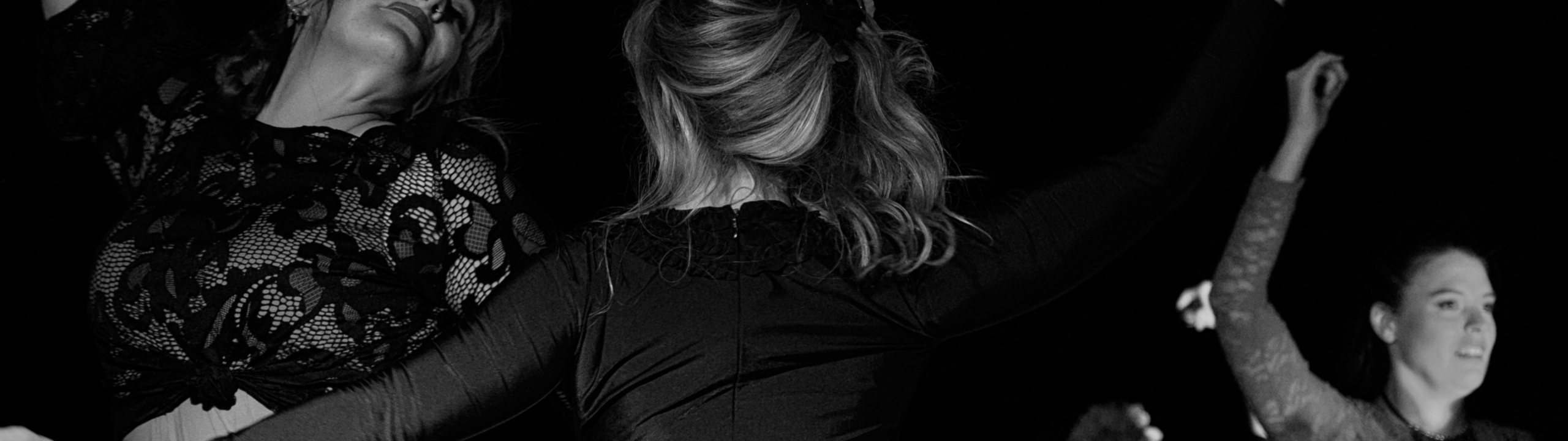 Three women dance joyfully, wearing dark outfits, with their arms raised. The photo is in black and white and captures their movement against a dark background.
