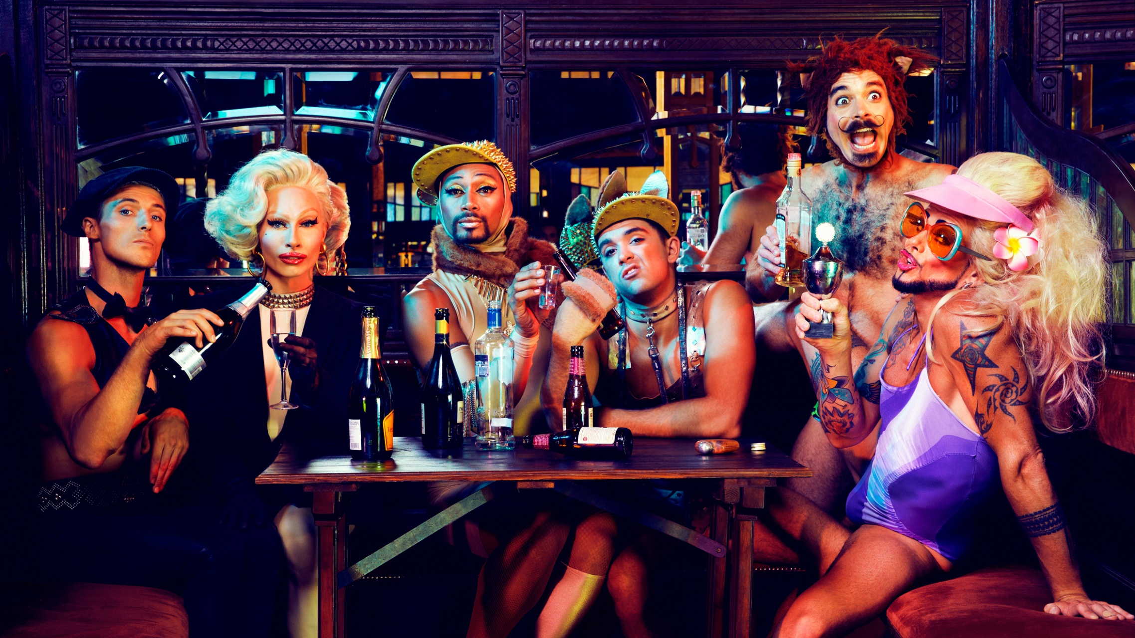 Six people in colorful, flamboyant outfits and makeup sit at a table with drinks, posing and smiling in a lively, festive atmosphere. Bottles and glasses are scattered on the table in front of them.