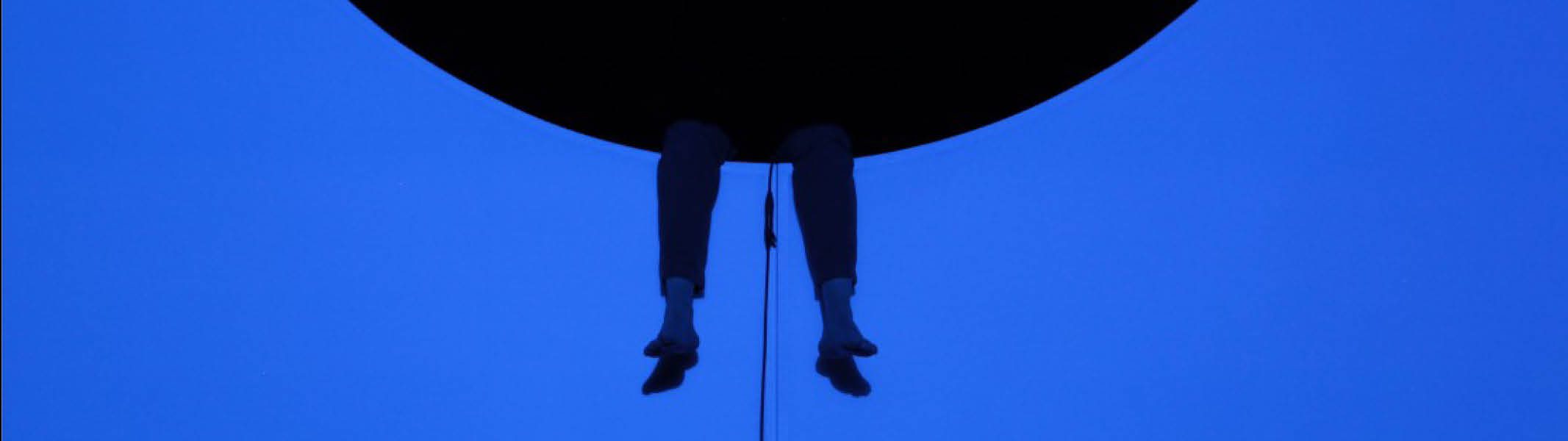 Silhouetted legs dangle from a large black circle above a bright blue background, creating a minimalist and surreal visual effect.