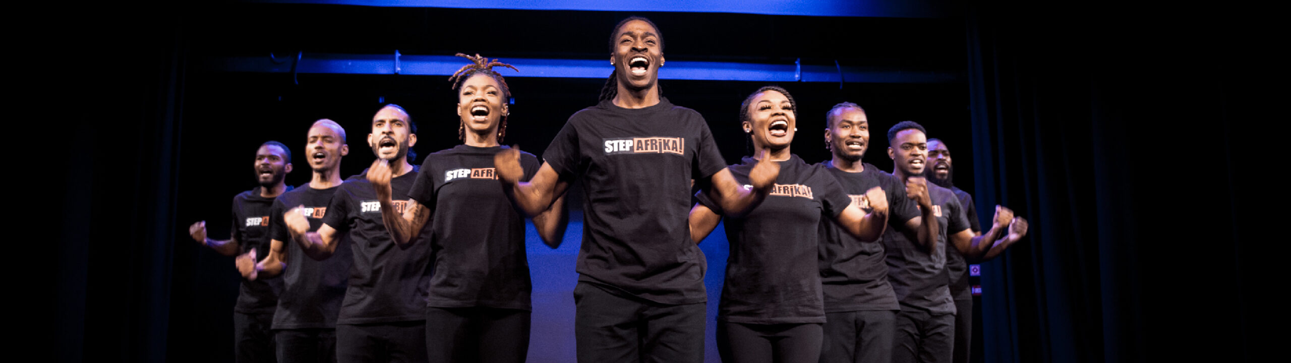 A group of performers in black “Step Afrika!” shirts stand in a line on stage, smiling and mid-performance, with bright stage lighting and a blue background.