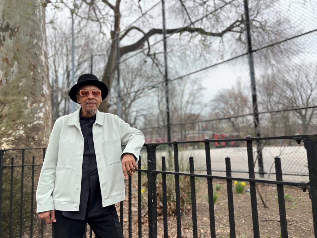 A man in a light jacket, black shirt, and hat stands by a black metal fence in front of a leafless tree and a fenced park area on a cloudy day.